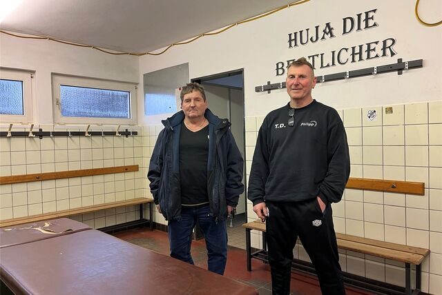 Ingo Reichel, Vereinsvorsitzender, und Thomas Droste, sportlicher Leiter beim SuS Bertlich, stehen in der Kabine des Vereins. Die Wände sind gekachelt, auf einer Kachel ist ein Aufkleber mit dem Vereinslogo befestigt, oben an der Wand steht ein Schriftzug.
