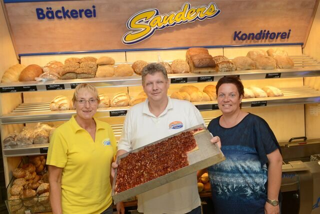 Theo Sanders, Sandra Sanders und eine Mitarbeiterin stehen vor dem Backregal. In der Hand hält er ein Blech mit Kuchen.