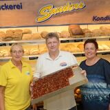 Theo Sanders, Sandra Sanders und eine Mitarbeiterin stehen vor dem Backregal. In der Hand hält er ein Blech mit Kuchen.