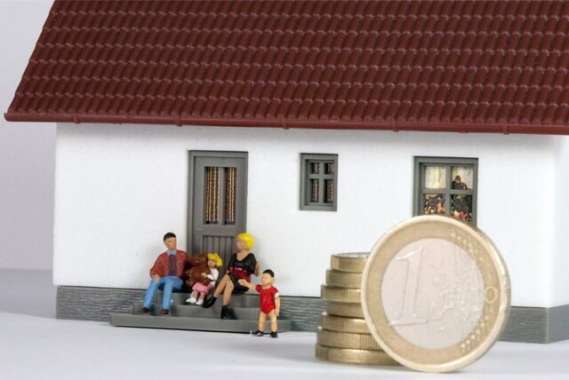Symbolbild zur Grundsteuer-Erhöhung: Kunststoff-Figuren, die  eine Familie darstellen, sitzen vor dem Modell eines Wohnhauses.