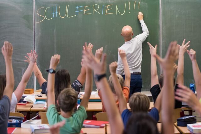 Ein Lehrer schreibt vor jubelnden Schülern „Schulferien“ an die Tafel.