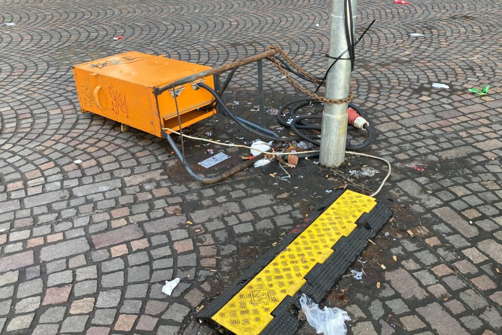 Auf dem Altstadtmarkt in Recklinghausen liegt ein orangefarbener Baustromkasten neben einem Laternenpfahl auf dem Pflaster.