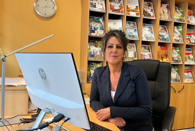 Eine Frau sitzt vor einer Katalogwand in einem Reisebüro am Schreibtisch, vor ihr ein Computerbildschirm.