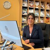 Eine Frau sitzt vor einer Katalogwand in einem Reisebüro am Schreibtisch, vor ihr ein Computerbildschirm.
