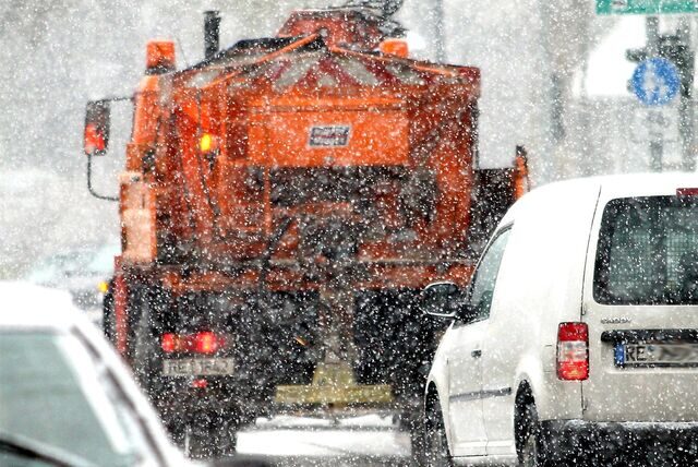 Ein oranges Streufahrzeug ist auf einer Straße unterwegs, es schneit.