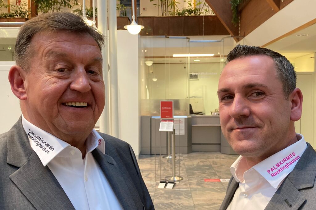 Zwei Männer stehen im Foyer der Sparkasse Vest Recklinghausen. Auf ihren Hemdkragen steht Schaustellerverein Recklinghausen und Palmkirmes Recklinghausen. Zwei Männer stehen im Foyer der Sparkasse Vest Recklinghausen. Auf ihren Hemdkragen steht Schaustellerverein Recklinghausen und Palmkirmes Recklinghausen.