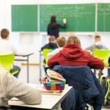 Eine Schulklasse von hinten. An der Tafel steht eine Lehrerin, die an die Tafel schreibt.