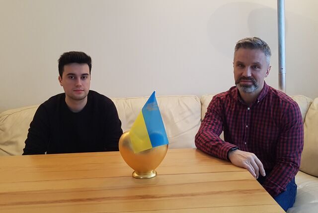 Ein junger Mann und ein Mann mittleren Alters sitzen auf einer Couch an einem Tisch in Recklinghausen. Zwischen ihnen steht eine Ukraine-Flagge