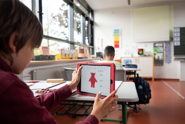 Ein Kind sitzt an einem Tisch in einem Klassenzimmer und hält eine Tablet hoch.