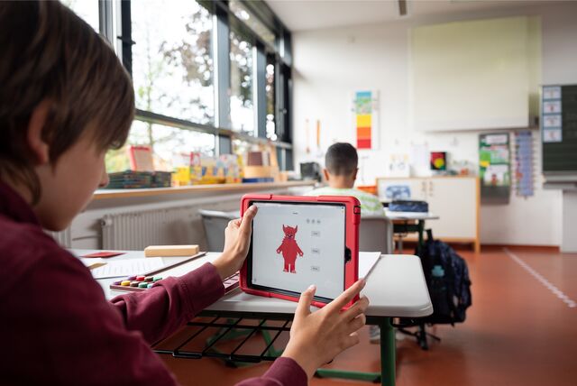 Ein Kind sitzt an einem Tisch in einem Klassenzimmer und hält eine Tablet hoch.