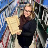 Kristina Schröder steht in einem engen Gang mit Regalen voller Aktenordner im Elisabeth-Krankenhaus in Recklinghausen und präsentiert ein altes Dokument.