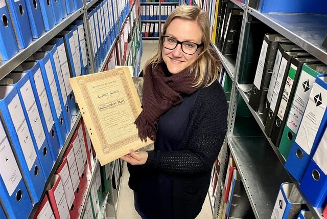 Kristina Schröder steht in einem engen Gang mit Regalen voller Aktenordner im Elisabeth-Krankenhaus in Recklinghausen und präsentiert ein altes Dokument.