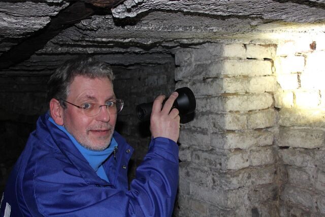 Stefan Wanske schaut in die Kamera und leuchtet gleichzeitig mit einer Taschenlampe auf eine gemauerte Wand.