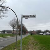 Im Vordergrund ist ein Straßenschild der Bestener Straße zu sehen, im Hintergrund die Straße selbst.