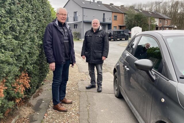 Zwei Senioren stehen zwischen einem Auto und einer Hecke auf einem Fußweg.