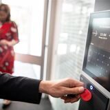 Eine Hand hält einen Chip an eine digitale Stempeluhr, im Hintergrund erkennt man eine Frau.