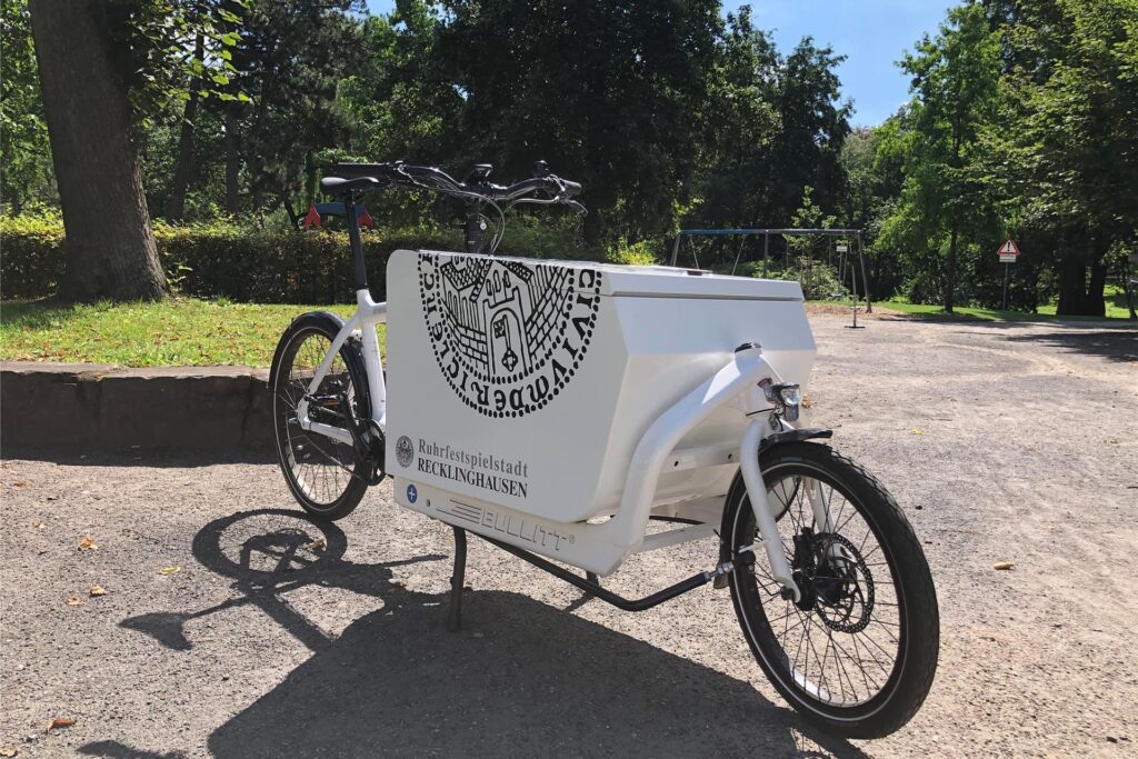 Ein weißes Lastenrad mit dem schwarzen Logo und Schriftzug der Ruhrfestspielstadt Recklinghausen steht an einem sonnigen Tag in einem Park. Ein weißes Lastenrad mit dem schwarzen Logo und Schriftzug der Ruhrfestspielstadt Recklinghausen steht an einem sonnigen Tag in einem Park.