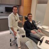 Dr. David Bonsmann sitzt auf einem Hocker, vor ihm im Behandlungsstuhl sitzt sein Patient Stefan Schlüter. Beide lächeln.