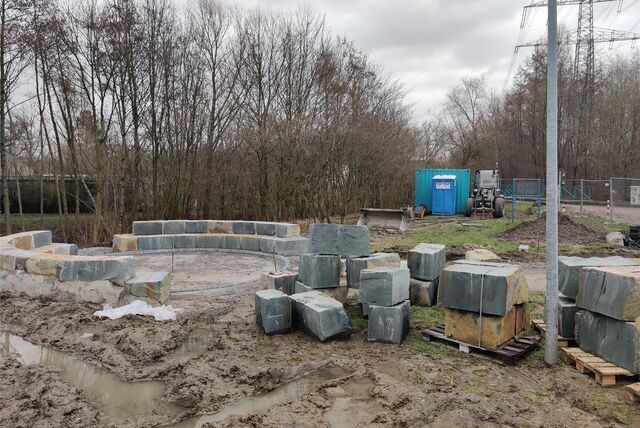 Zu sehen ist die Baustelle an der Nierholzstraße, einige Steinblöcke sind bereits ringförmig aufgestellt. Das restliche Material steht aufgehäuft oder auf Paletten, aufgrund des Regenfalls ist der Boden matschig.