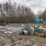 Zu sehen ist die Baustelle an der Nierholzstraße, einige Steinblöcke sind bereits ringförmig aufgestellt. Das restliche Material steht aufgehäuft oder auf Paletten, aufgrund des Regenfalls ist der Boden matschig.
