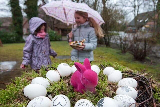 Zwei Mädchen stehen mit Regenschirm in einem Garten. Ein Mädchen hält ein buntes Osternest in der Hand, im Vordergrund steht ebenfalls ein Nest mit weißen Eiern und einer rosa Hasenfigur.