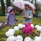 Zwei Mädchen stehen mit Regenschirm in einem Garten. Ein Mädchen hält ein buntes Osternest in der Hand, im Vordergrund steht ebenfalls ein Nest mit weißen Eiern und einer rosa Hasenfigur.