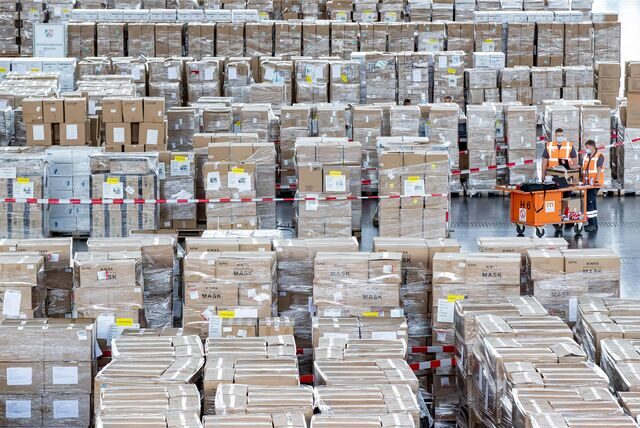 Mitarbeiter mit Schutzmasken arbeiten zwischen zahlreichen Kartons mit Schutzausrüstung in Millionenzahl.