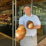 Ein Mann in weißem Polohemd und Schürze steht vor einer Bäckereifiliale in Waltrop. In den Händen hält er zwei Brote. (Archivbild)