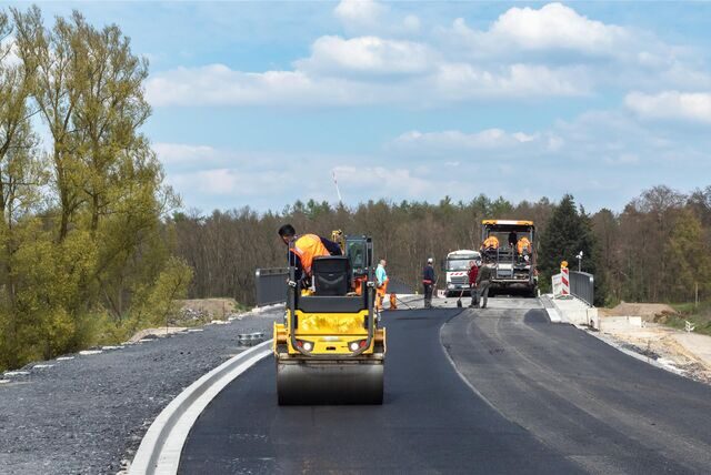 Eine kleine Walze fährt über frisch aufgebrachten Straßenbelag. Im hinteren Bereich des Bildes ist ein Asphaltfertiger zu sehen.