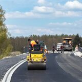 Eine kleine Walze fährt über frisch aufgebrachten Straßenbelag. Im hinteren Bereich des Bildes ist ein Asphaltfertiger zu sehen.