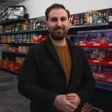 Ein Mann steht in einem Innenraum, hinter ihm steht eine Eismaschine, Regale mit Getränken, Lebensmitteln und Süßigkeiten.