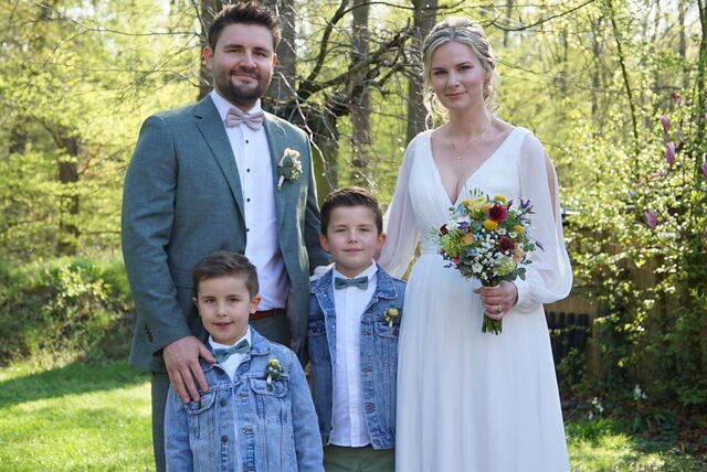 Ein Mann im Anzug und eine Frau im weißen Kleid stehen gemeinsam mit zwei Jungen auf einer Wiese.
