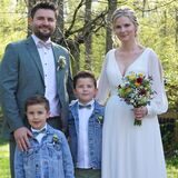 Ein Mann im Anzug und eine Frau im weißen Kleid stehen gemeinsam mit zwei Jungen auf einer Wiese.