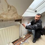 Ibrahim Gören hockt auf dem Boden seines Schlafzimmers. Er zieht ein Stück Tapete von der Wand, große grau-gelbe Flecken sind zudem zu erkennen.