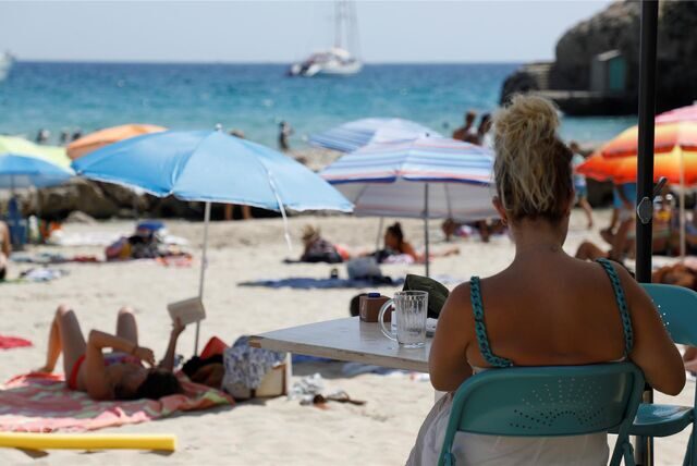 Eine Frau sitzt an einem Strand, im Hintergrund sind viele Menschen mit Sonnenschirmen zu sehen.