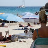 Eine Frau sitzt an einem Strand, im Hintergrund sind viele Menschen mit Sonnenschirmen zu sehen.