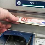 Eine Person zieht mit der linken Hand einige Geldscheine aus dem Schlitz eines Geldautomaten.