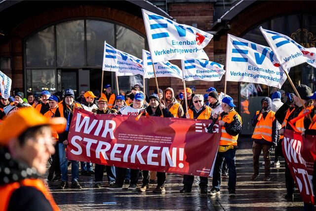 Demonstranten der Eisenbahn- und Verkehrsgewerkschaft (EVG) stehen mit Plakaten vor einem Hauptbahnhof.