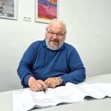 Wolfgang Bollrath sitzt an einem weißen Tisch und zeigt mit einem Stift auf den fertigen Entwurf.
