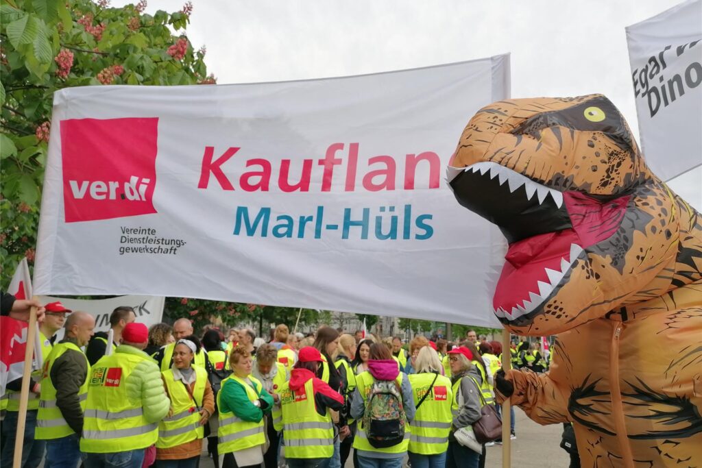 Zu sehen ist eine Person in T-Rex Kostüm die ein Schild mit der Aufschrift 