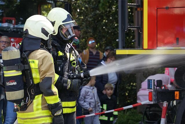 Zwei Feuerwehrleute in Uniform und Atemschutzmaske demonstrieren eine Löschübung. Aus dem Schlauch spritzt Wasser.