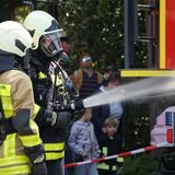 Zwei Feuerwehrleute in Uniform und Atemschutzmaske demonstrieren eine Löschübung. Aus dem Schlauch spritzt Wasser.