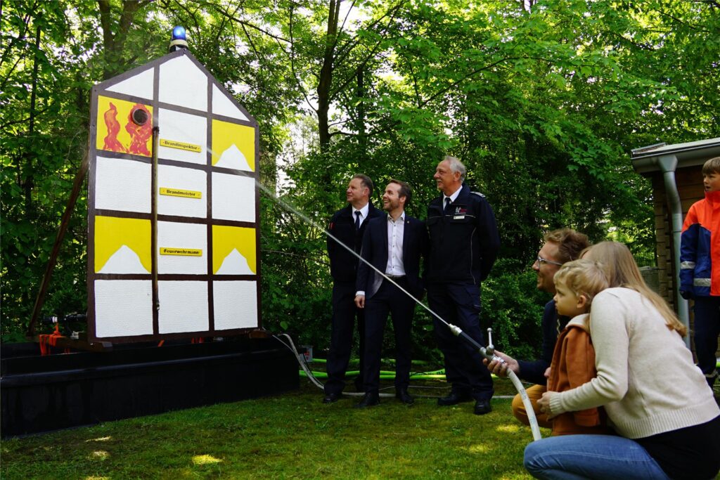 Ein Kind und seine Eltern hocken vor der Spritzwand, im Hintergrund stehen zwei Feuerwehrleute und der Waltroper Bürgermeister.