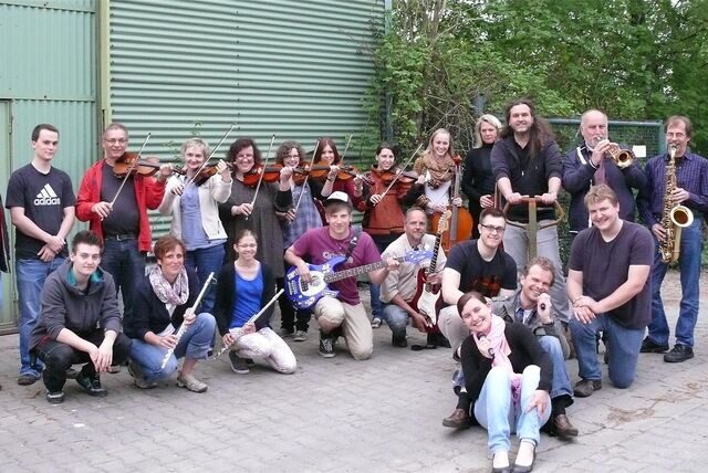 Menschen mit Musikinstrumenten stehen und hocken vor einer Lagerhalle.