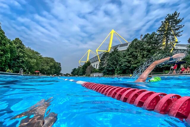 Eine Person schwimmt in einem Schwimmbecken des Freibads Volkspark in Dortmund. Im Hintergrund ist der Signal Iduna Park zusehen.