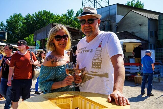 Ein Paar (Mann und Frau mit Sonnenbrillen und vollen Weingläsern in der Hand) steht an einem Bistrotisch.