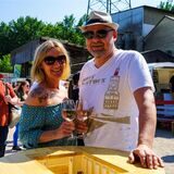Ein Paar (Mann und Frau mit Sonnenbrillen und vollen Weingläsern in der Hand) steht an einem Bistrotisch.