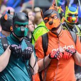Drei Männer in bunten T-Shirts und mit abstrakten Hunde-Masken im Gesicht.