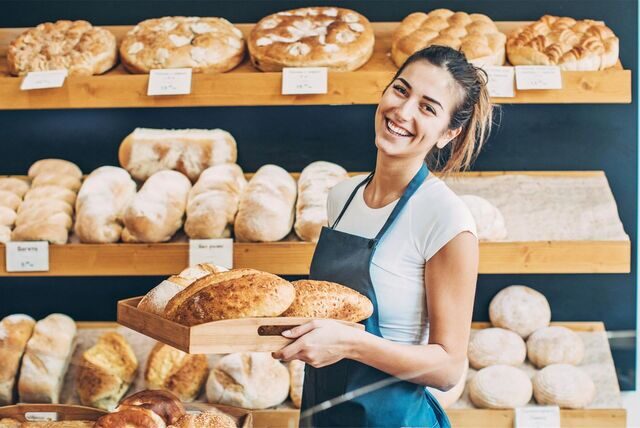 Eine Mitarbeiterin einer Bäckerei steht vorm Verkaufsregal und hält ein Tablet mit Brot in der Hand.
