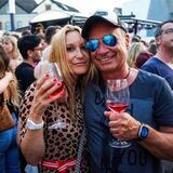 Ein Mann und eine Frau schauen mit Weingläsern in der Hand in die Kamera. Im Hintergrund sind viele weitere Menschen zu sehen.